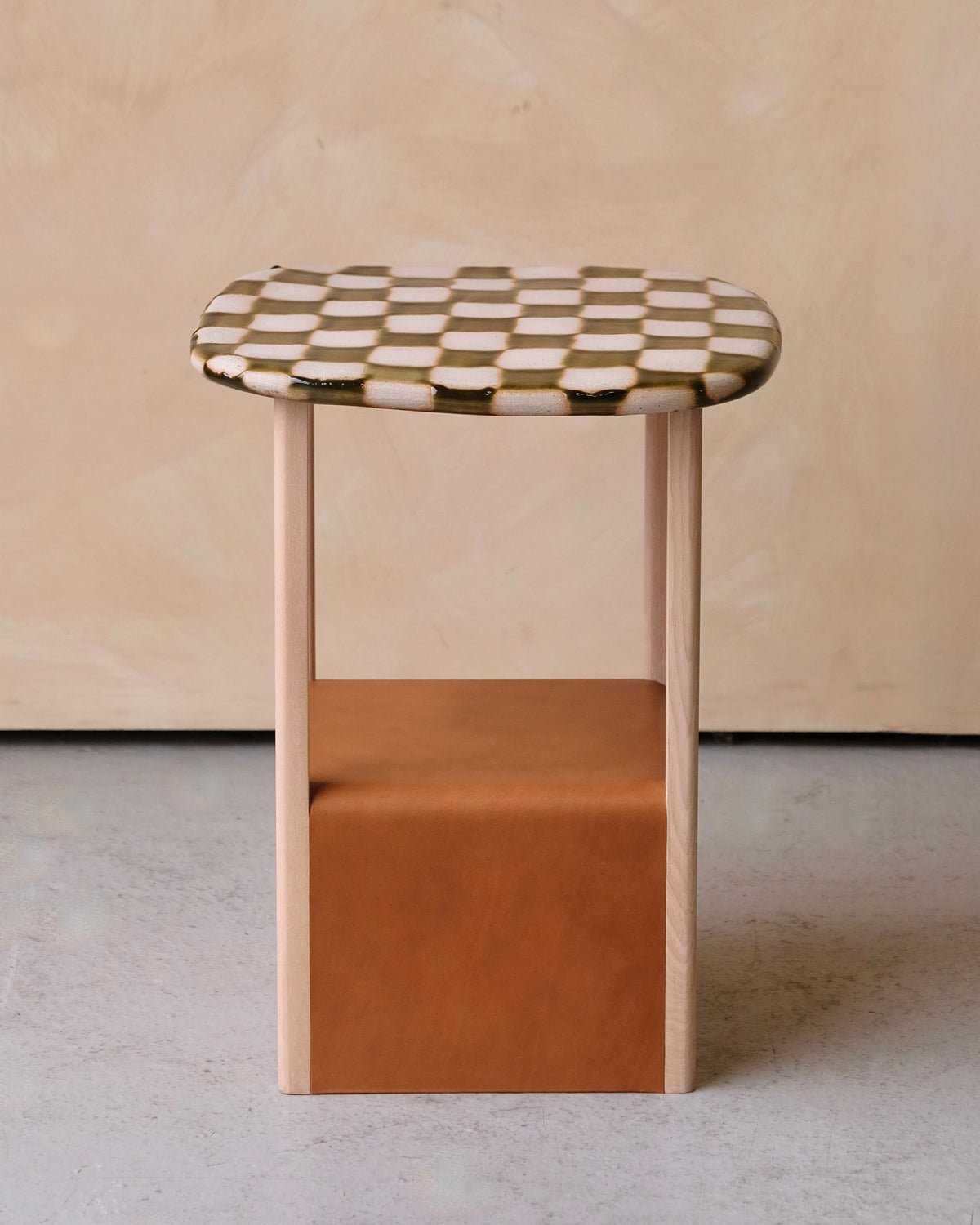 A small wooden side table with a checkered top in cream and dark brown, light natural wood legs, and a tan leather storage cube in the middle, set against a textured beige wall and concrete floor.