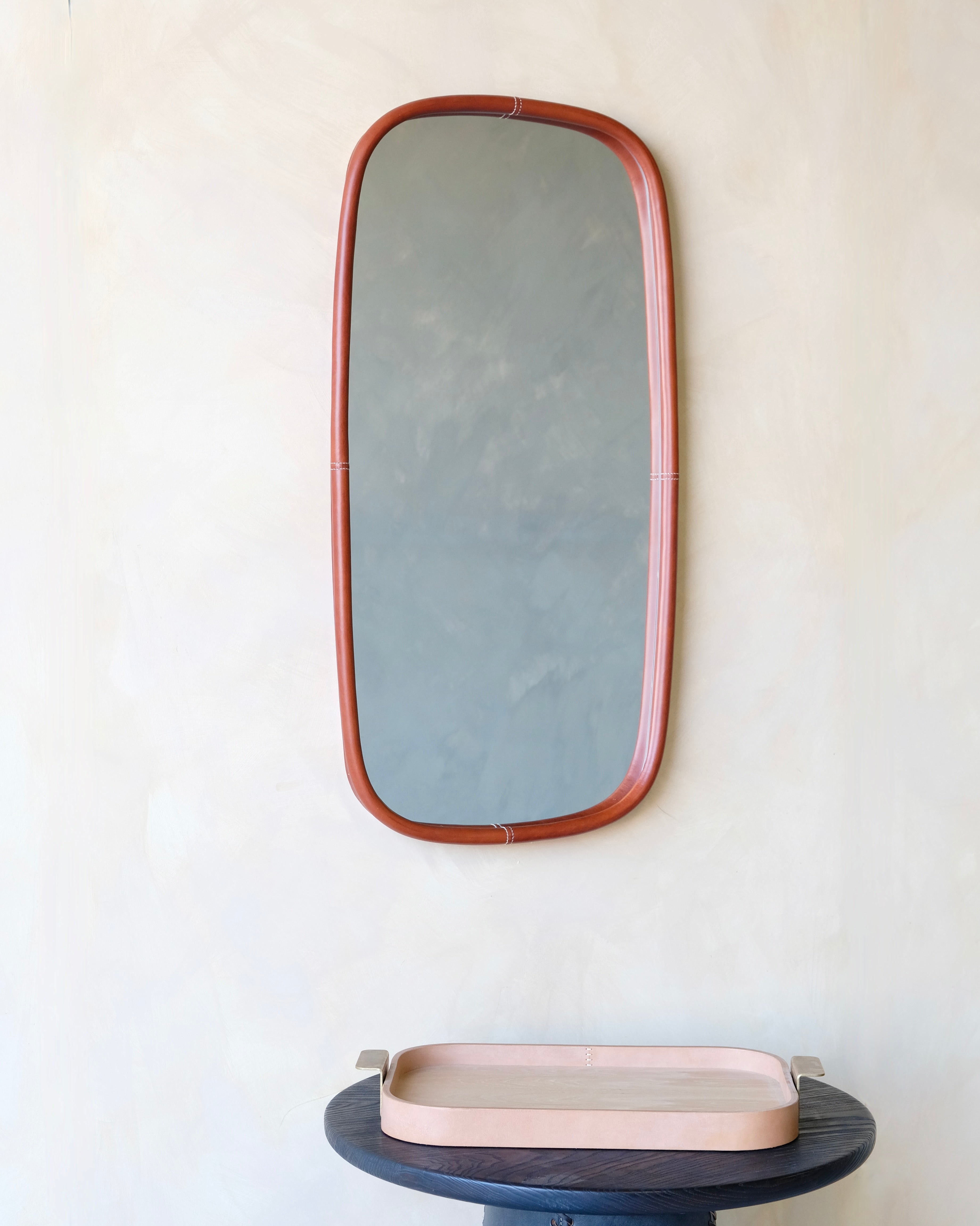 Rectangular mirror with leather frame on a white wall above a small round table.